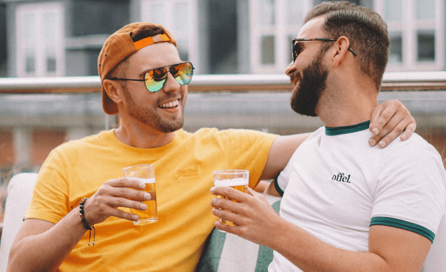 Two men face each other with an arm around each other and a beer in each hand