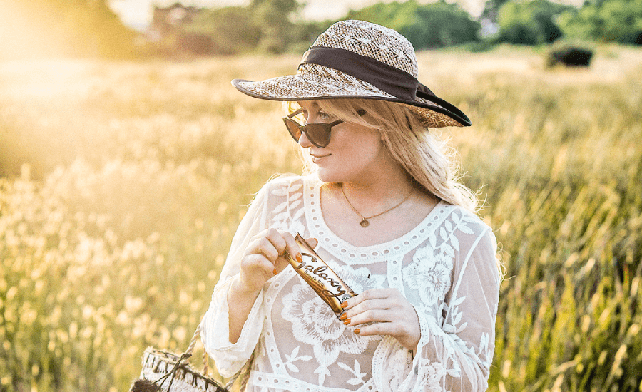 Woman wearing a hat, sunglasses and white lace holds a chocolate bar and looks into the distance in front of a field